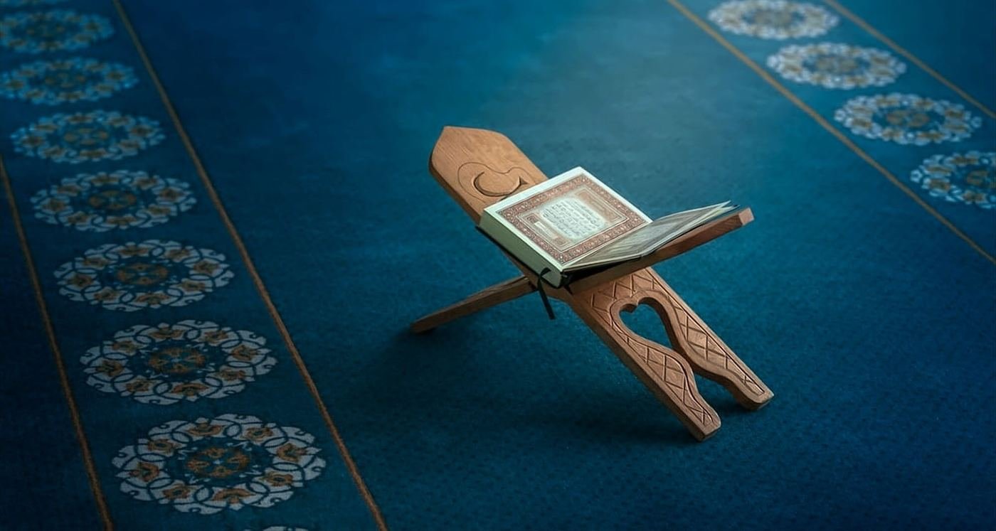 An open Qur'an resting on a wooden rehal on a blue mosque carpet
